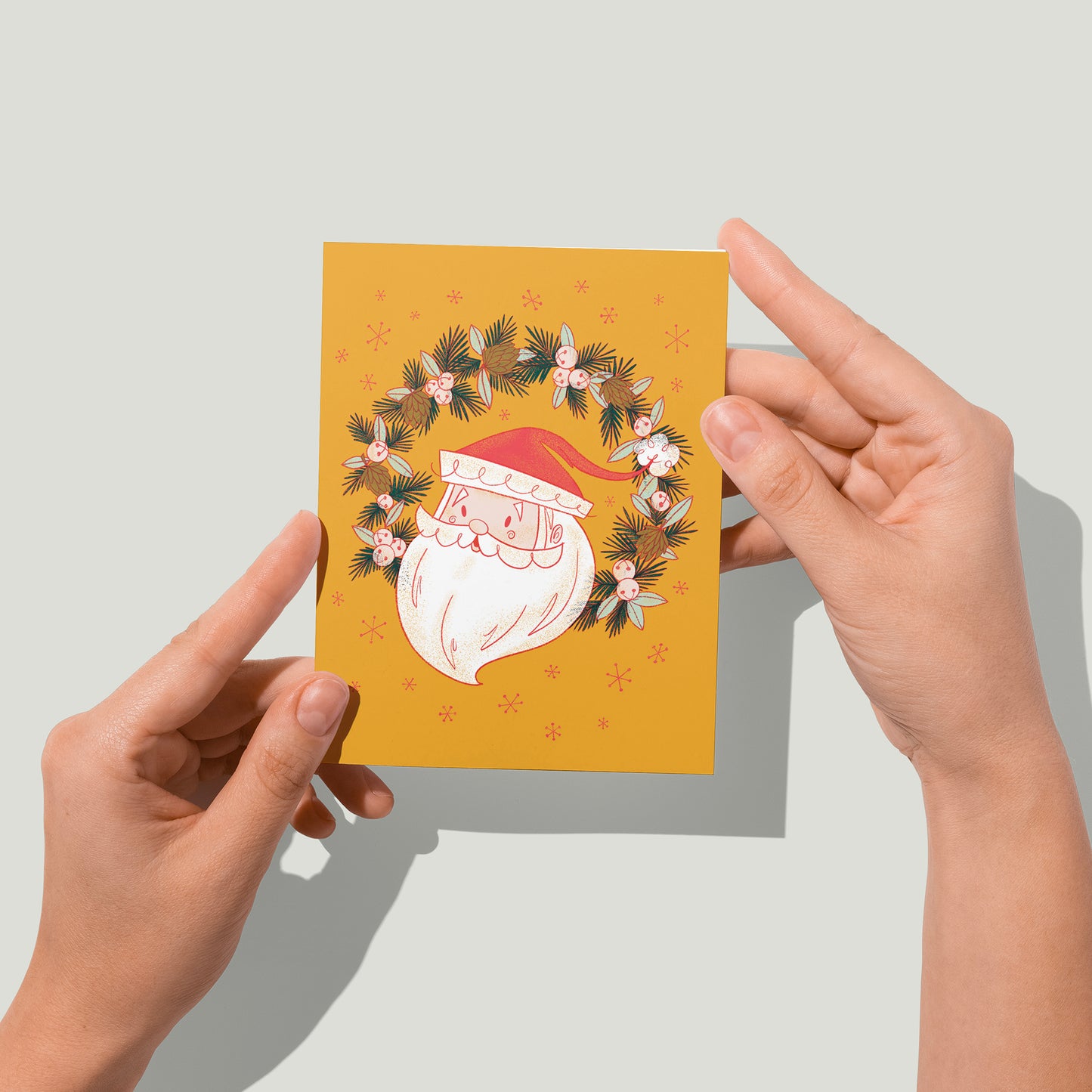 A yellow card shows a midcentury-style illustration of Santa's head in a wreath of jingle bells, pinecones and evergreen branches. A pair of hands holds the card up in front of the camera.