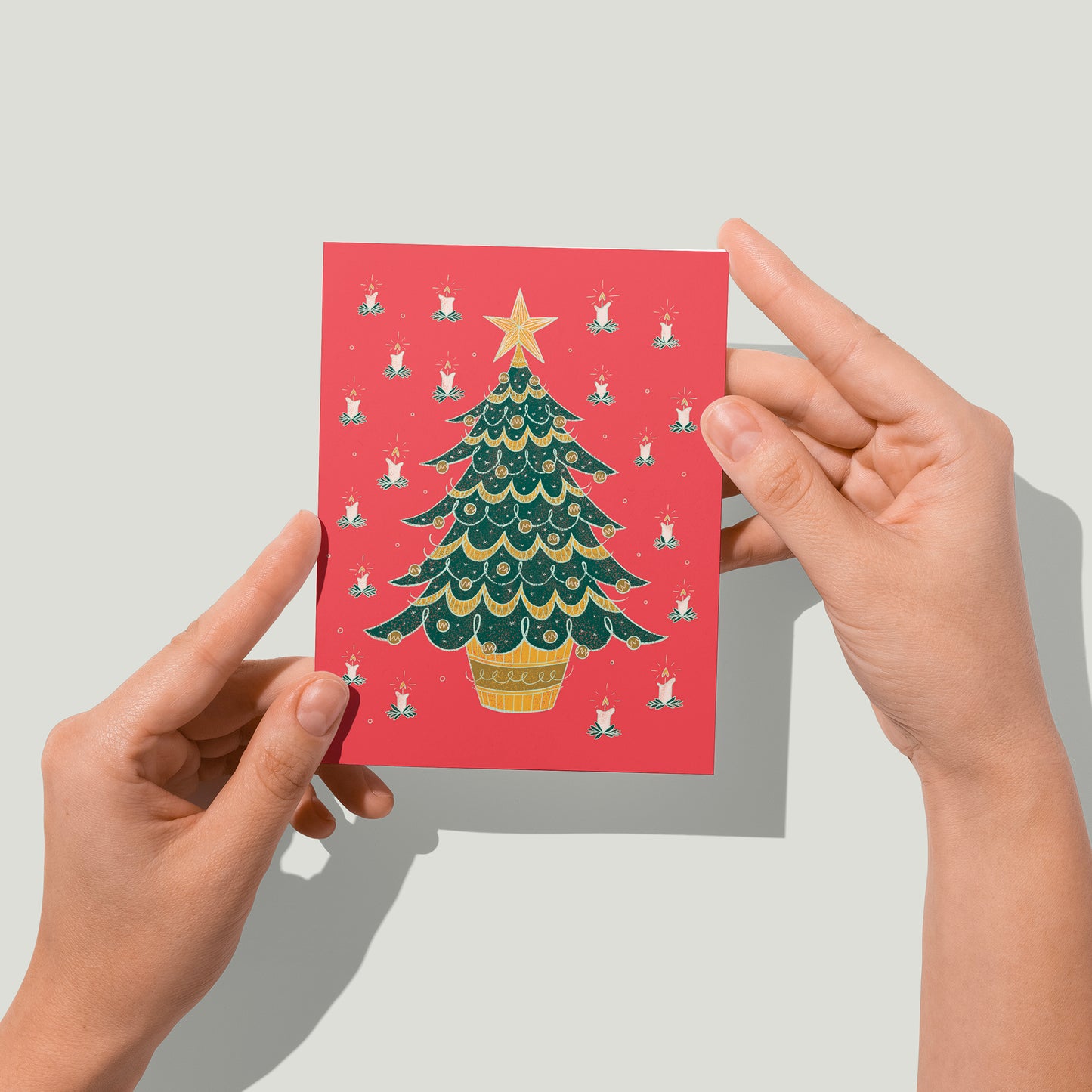 A red card features a midcentury-style bushy evergreen tree in a pot covered in baubles and garland, around which is a pattern of lit candles. A pair of hands holds the card up in front of the camera.