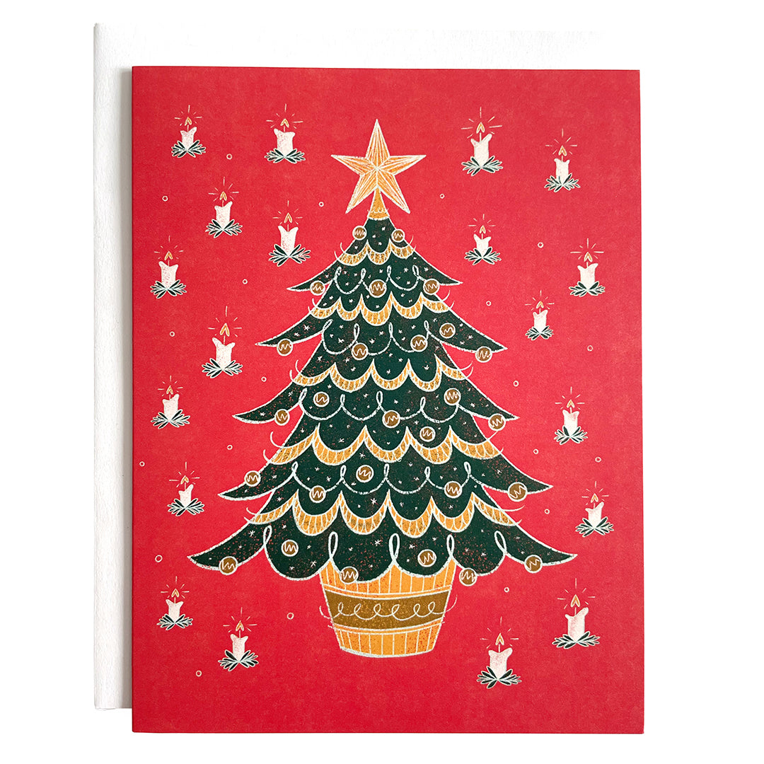 A red card features a midcentury-style bushy evergreen tree in a pot covered in baubles and garland, around which is a pattern of lit candles. The card is against a white envelope on a white background.