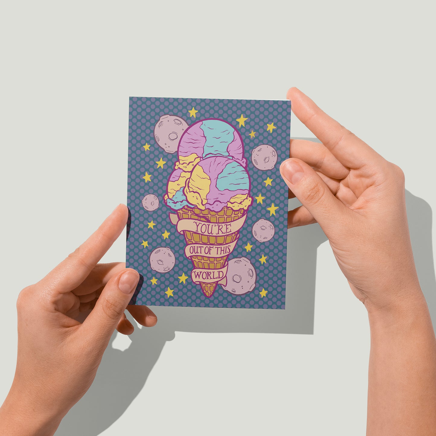 A purple greeting card features a triple scoop waffle cone of Moon Mist ice cream, purple moons, yellow stars and a banner that reads you're out of this world in hand lettering. A pair of hands holds the card up in front of the camera.