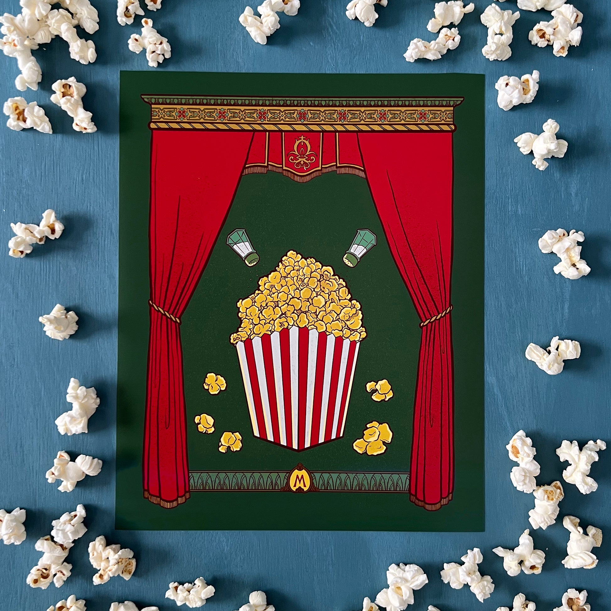 This art print shows a full box of buttery popcorn with salt shakers above it, framed by rich curtains and a detailed capital. An M monogram appears at the bottom. The print is shown unframed and surrounded by popcorn on a blue background.