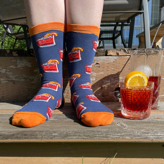 A person wears blue and orange socks patterned with negronis garnished with orange wedges. They stand on a step on a deck next to a negroni in a double old-fashioned glass and a mixing glass with more negroni in it.