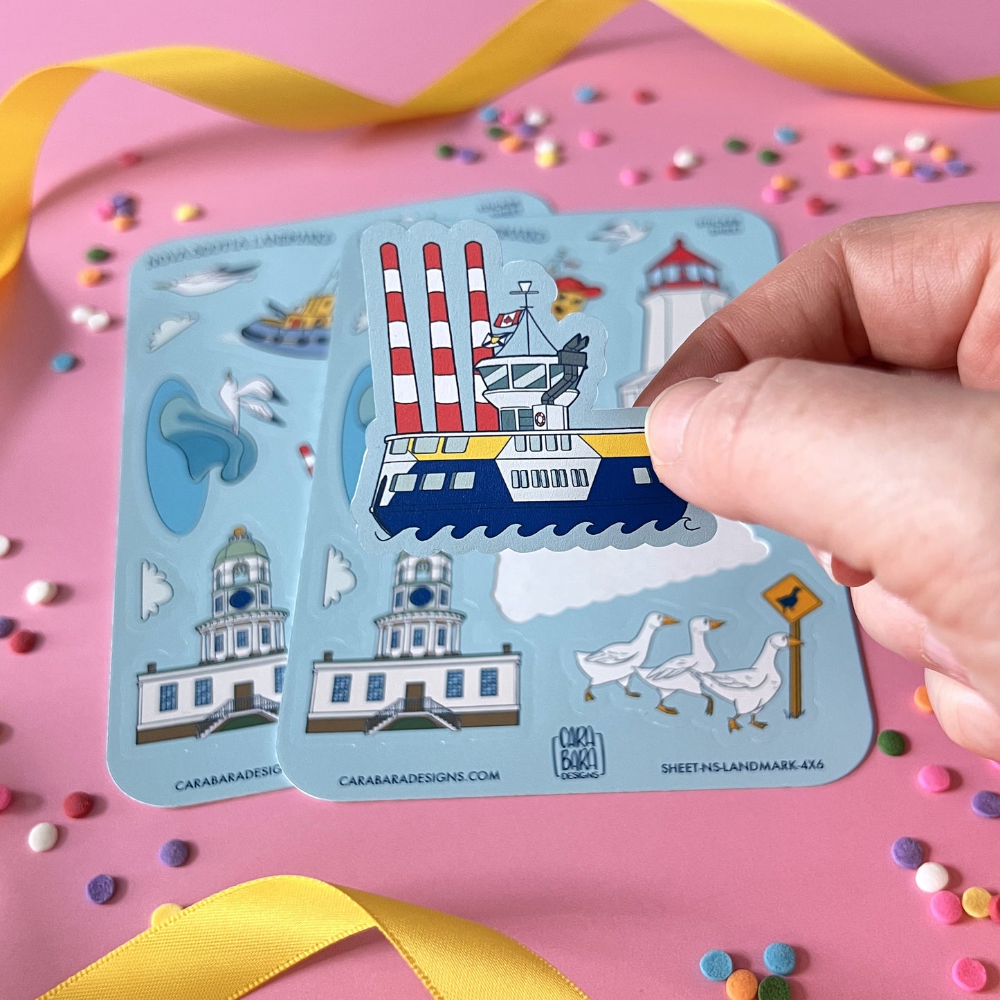 Two blue vinyl sticker sheets featuring landmarks from Halifax and in and around Nova Scotia are on a pink background with confetti and ribbon. A hand holds up a sticker of the Dartmouth ferry to the camera.