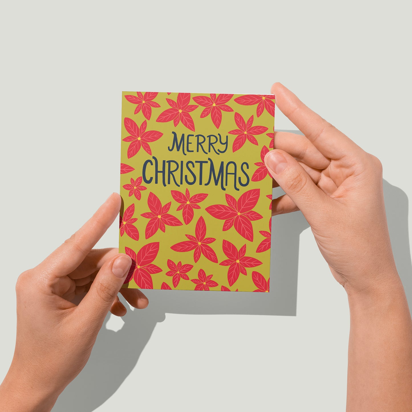 A bright green card features a pattern of bright red poinsettias and the words Merry Christmas in navy in the upper third. A pair of hands holds the card up in front of the camera.