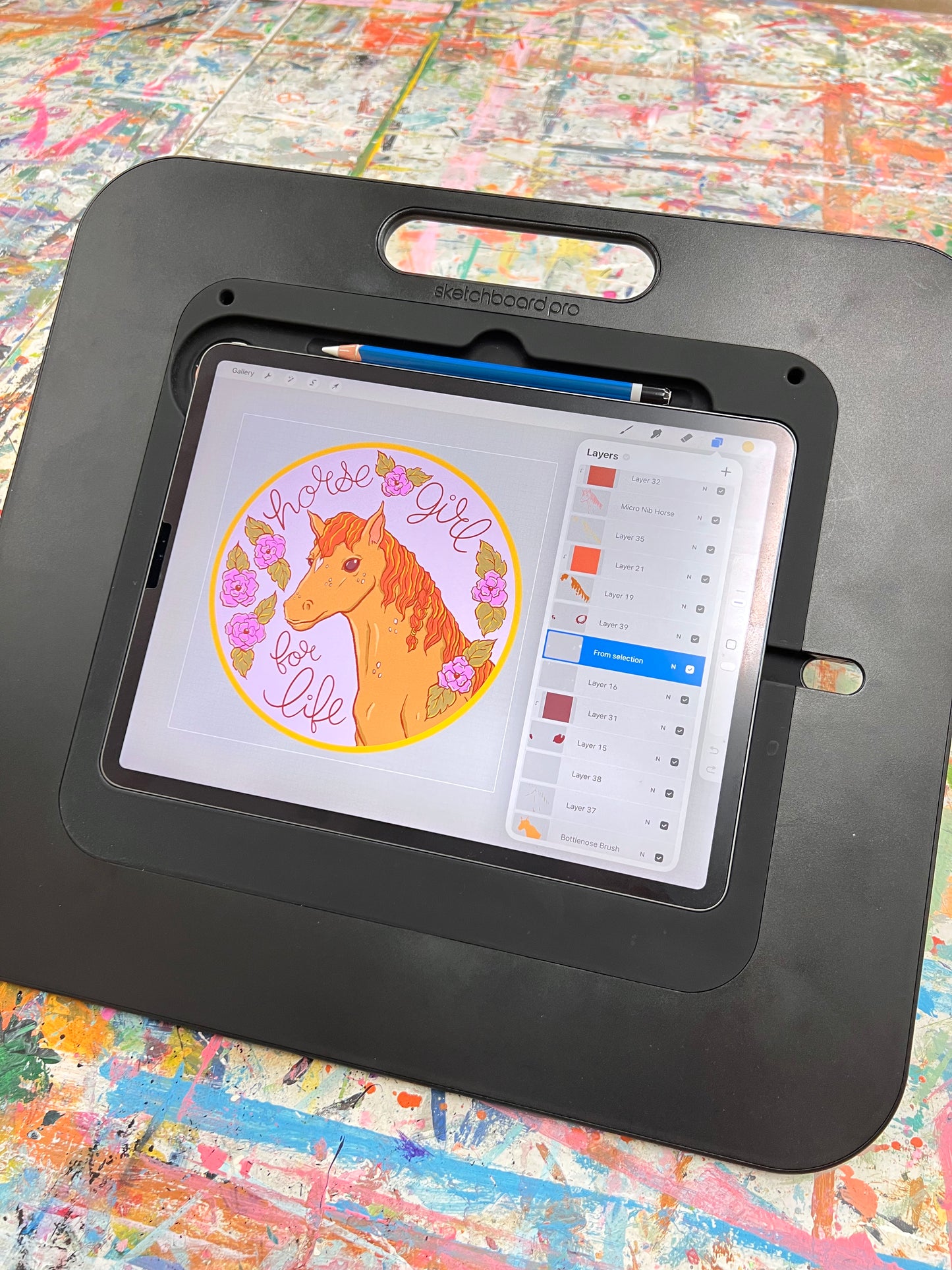 An ipad is set up on a paint-splattered table, open to a sticker design in the procreate app with the layer menu open.