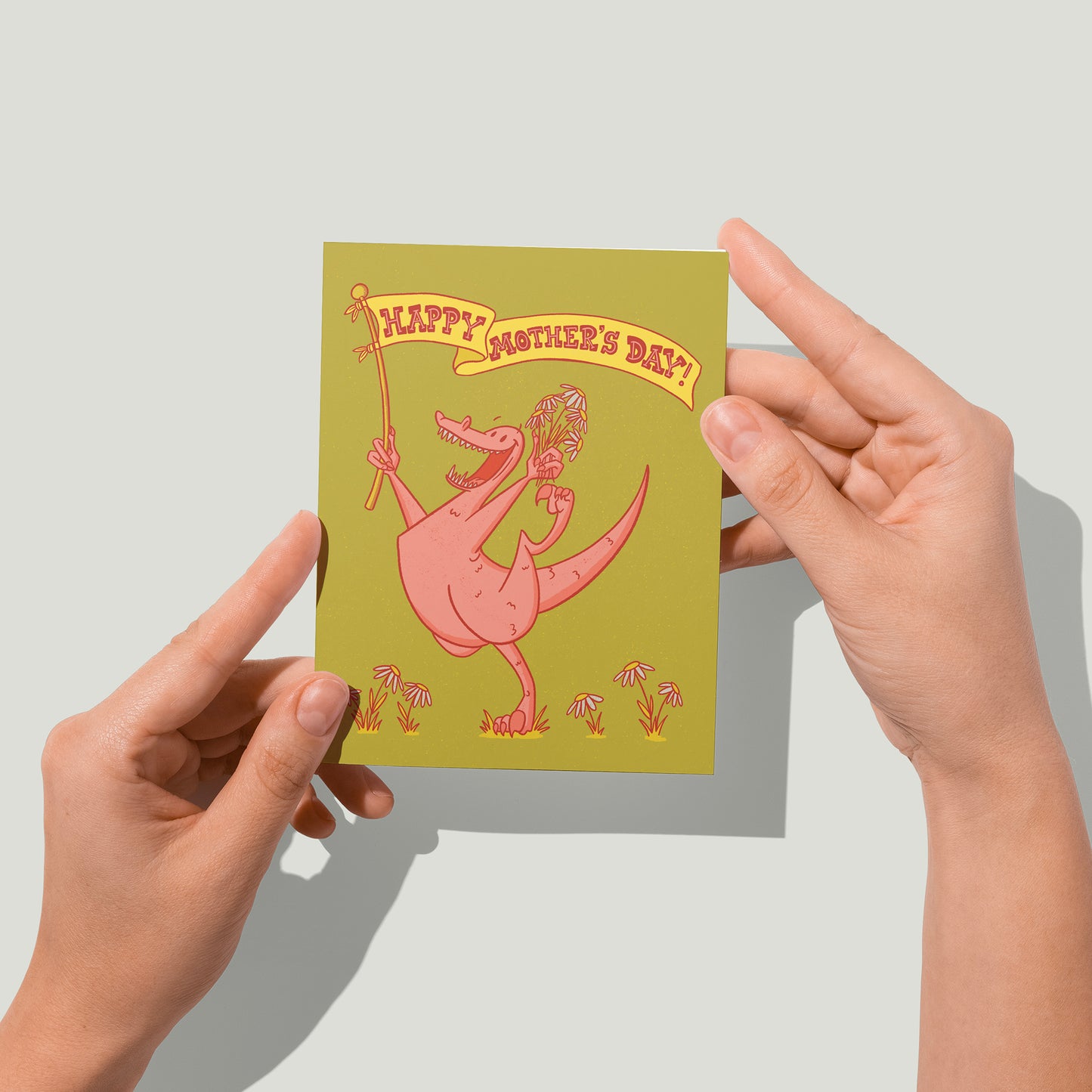 A green card shows an exuberant pink dinosaur has a handful of daisies and a hand-lettered yellow banner that reads Happy Mother's Day. A pair of hands holds the card up in front of the camera.