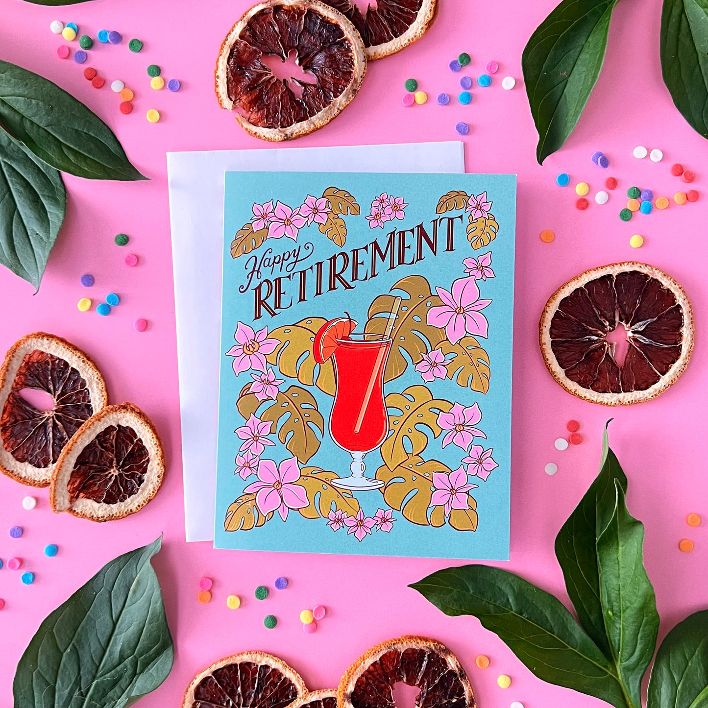 A blue retirement card with a hurricane cocktail, orchids and leaves is on pink with dried citrus, confetti, and leaves.