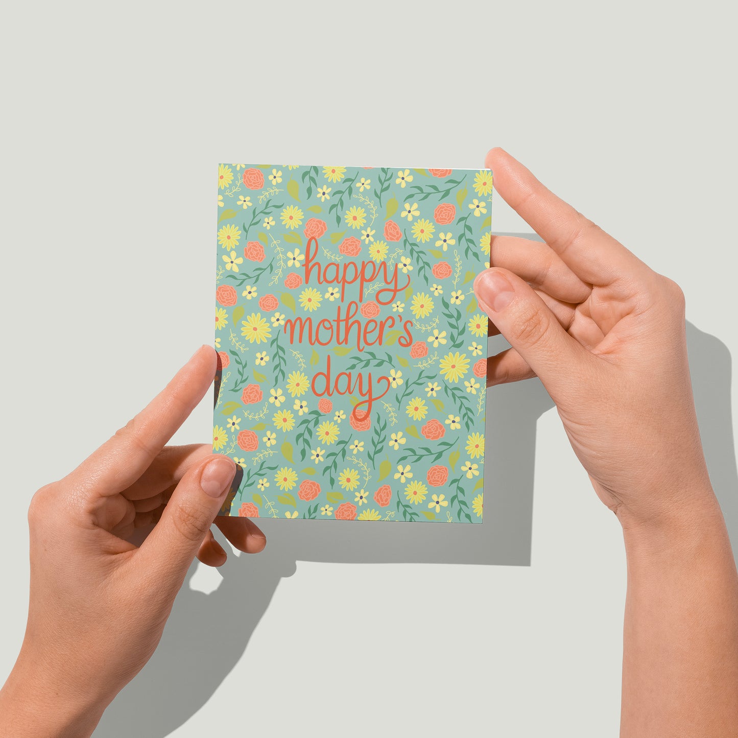 A mint-green greeting card is covered with a pattern of tiny yellow and pink flowers and the words happy mother's day in red letters. A pair of hands holds the card up in front of the camera.