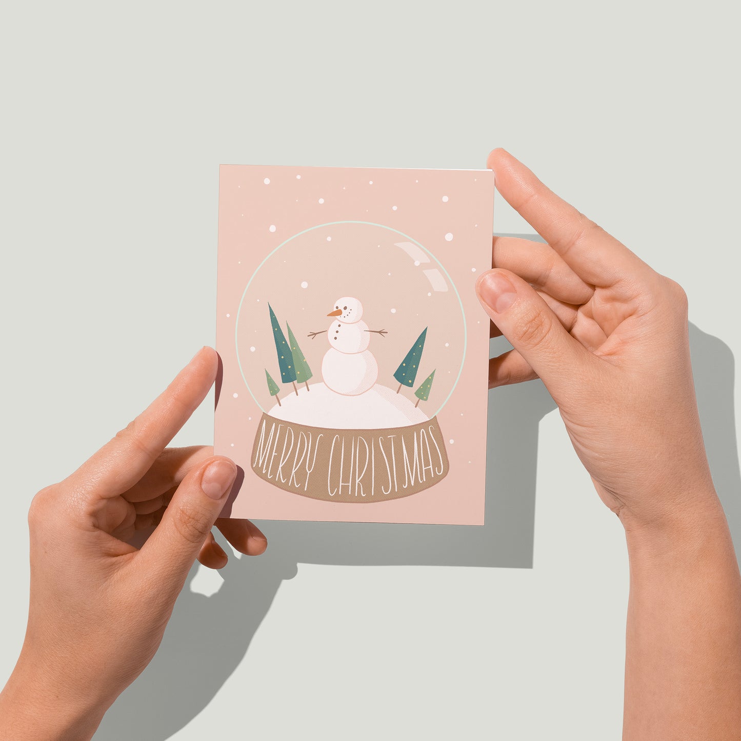 A pink Christmas card features an illustration of a snow globe with a cheerful snowman, triangular evergreens, and the words Merry Christmas lettered at its base. A pair of hands holds the card up in front of the camera.