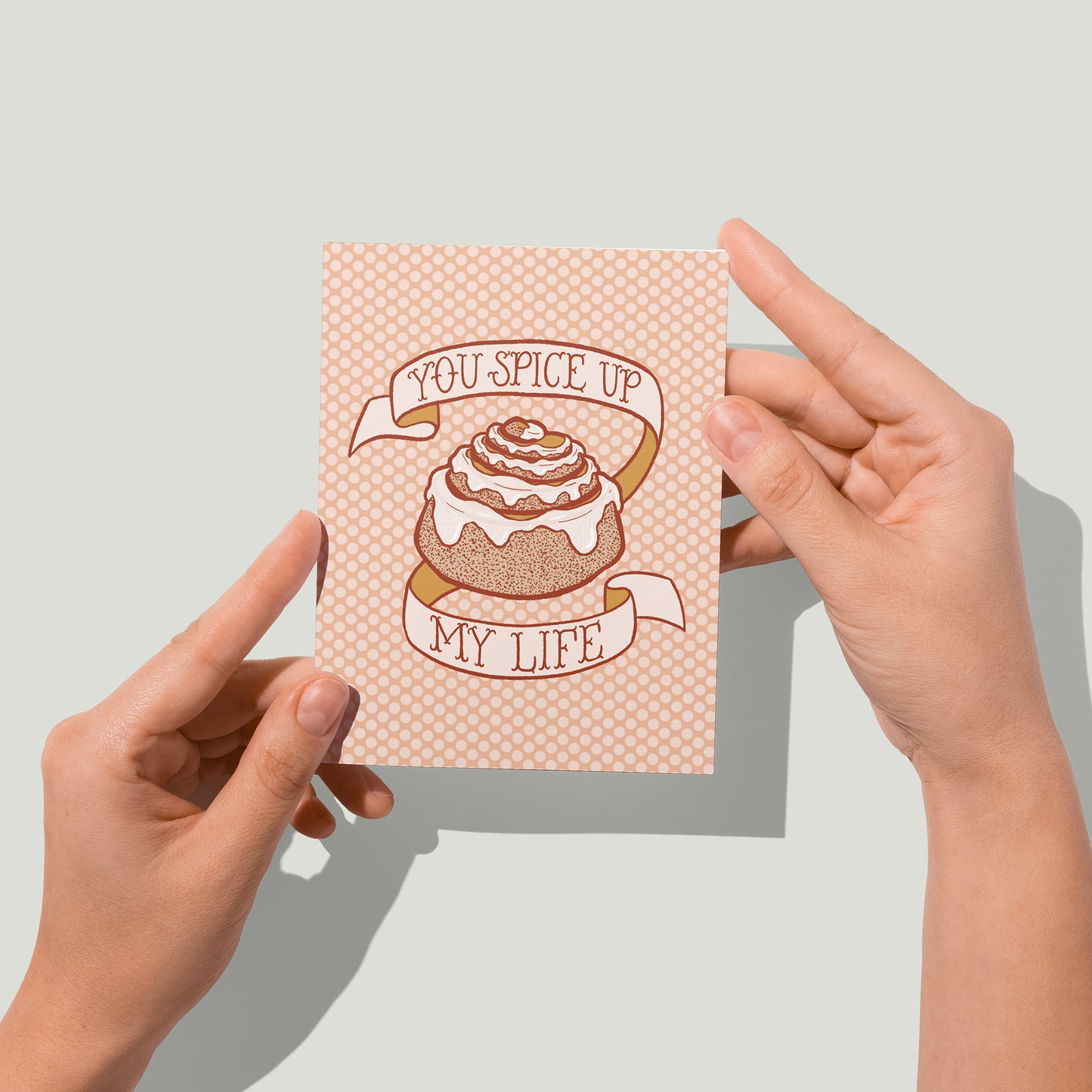 A peach polka dot greeting card bears an illustration of a cinnamon bun dripping with frosting and a banner that reads you spice up my life in hand lettering. A pair of hands holds the card up in front of the camera.