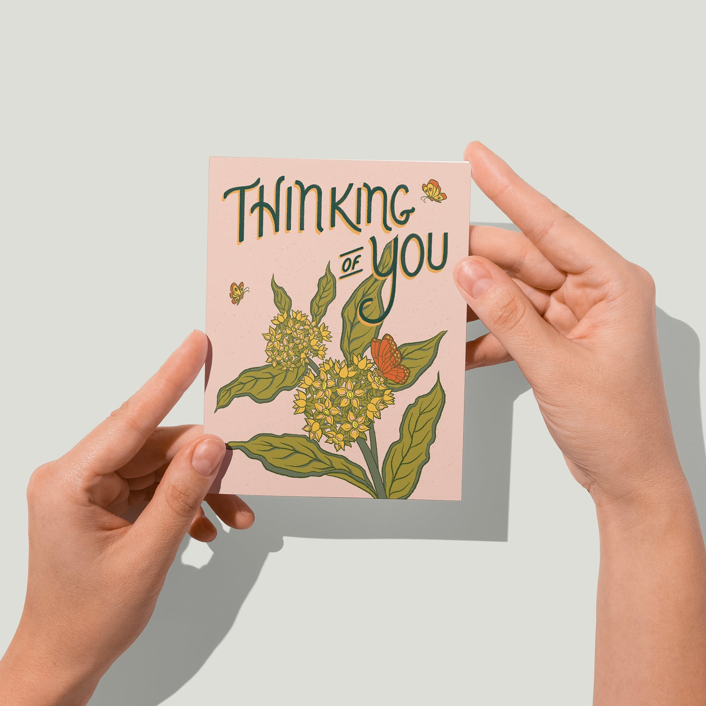 A pale pink greeting card has the words Thinking of You in dark green with a burst of milkweed blossoms and monarch butterflies below the letters. A pair of hands holds the card up in front of the camera.