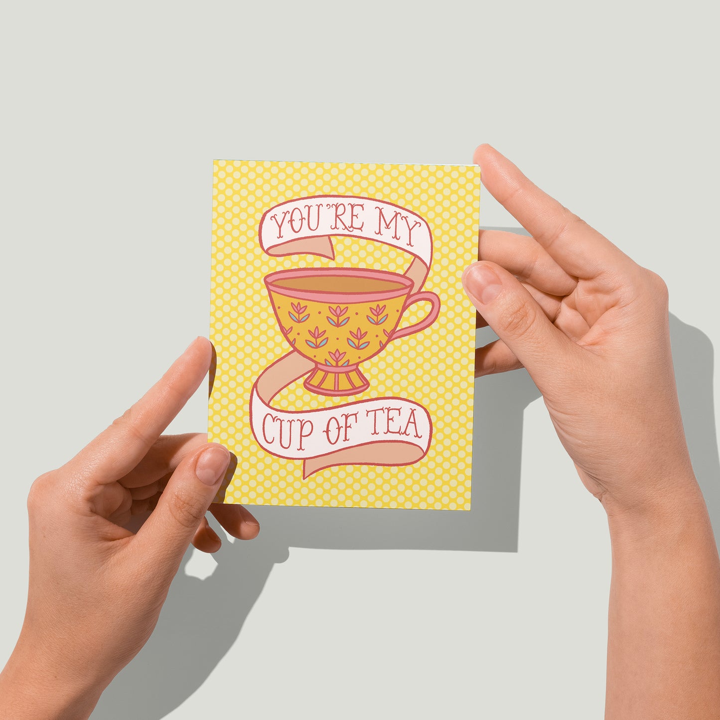 A yellow polka dot greeting card features an illustration of a golden teacup with yellow flowers, full of strong tea. Around the teacup is a banner that reads You're my cup of tea in pink letters. A pair of hands holds the card up in front of the camera.