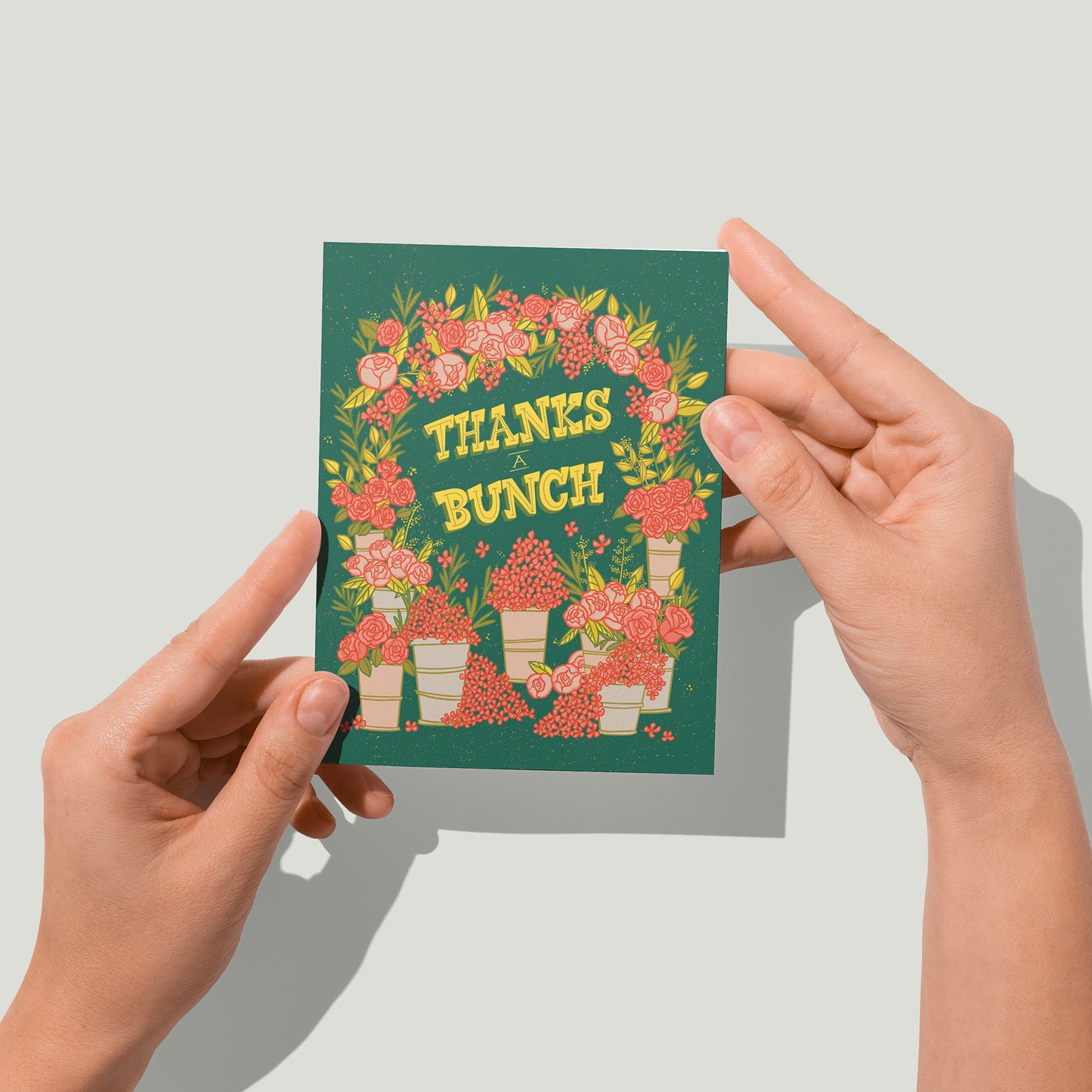 A dark green greeting card shows an arch covered in pink flowers with buckets of flowers at the bottom. It reads Thanks a Bunch in yellow letters. A pair of hands holds the card up in front of the camera.