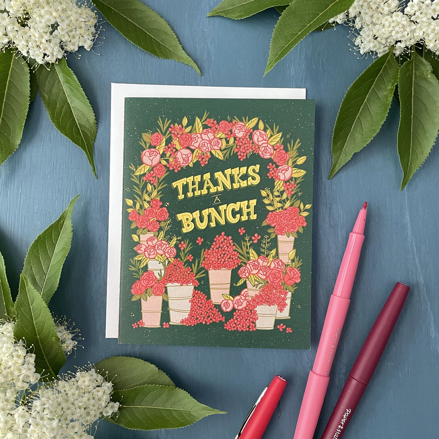 A dark green greeting card shows an arch covered in pink flowers with buckets of flowers at the bottom. It reads Thanks a Bunch in yellow letters. It is on a blue background flanked by white flowers, leaves and felt-tip pens.
