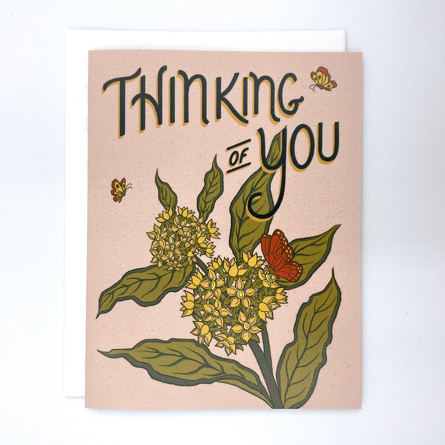 A pale pink greeting card has the words Thinking of You in dark green with a burst of milkweed blossoms and monarch butterflies below the letters. The card is against a white envelope on a white background.
