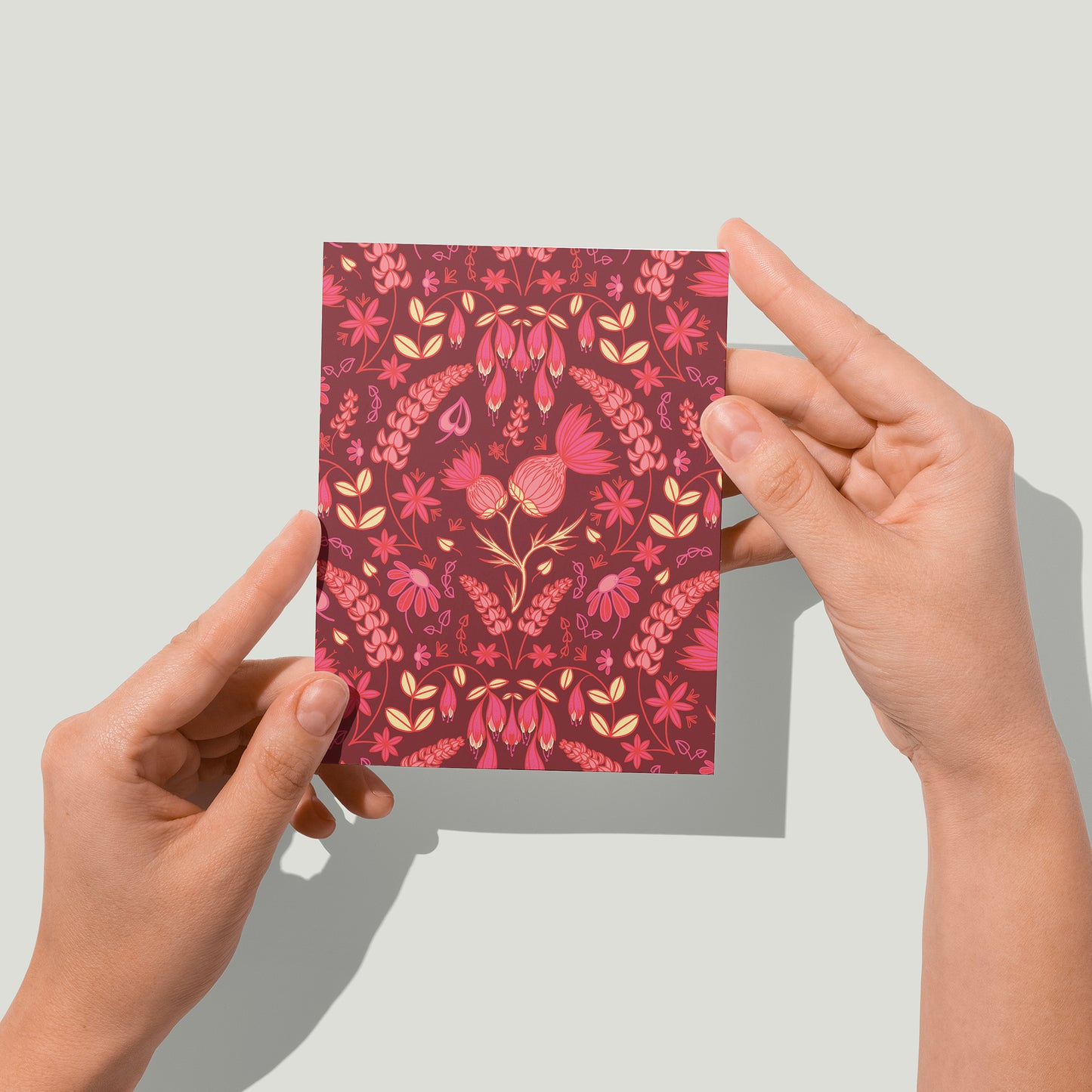 A dark pink greeting card is covered in a folk-style pattern of pink thistles, lupines, haskap berries and mayflowers, with creamy yellow leafy accents. A pair of hands holds the card up in front of the camera.