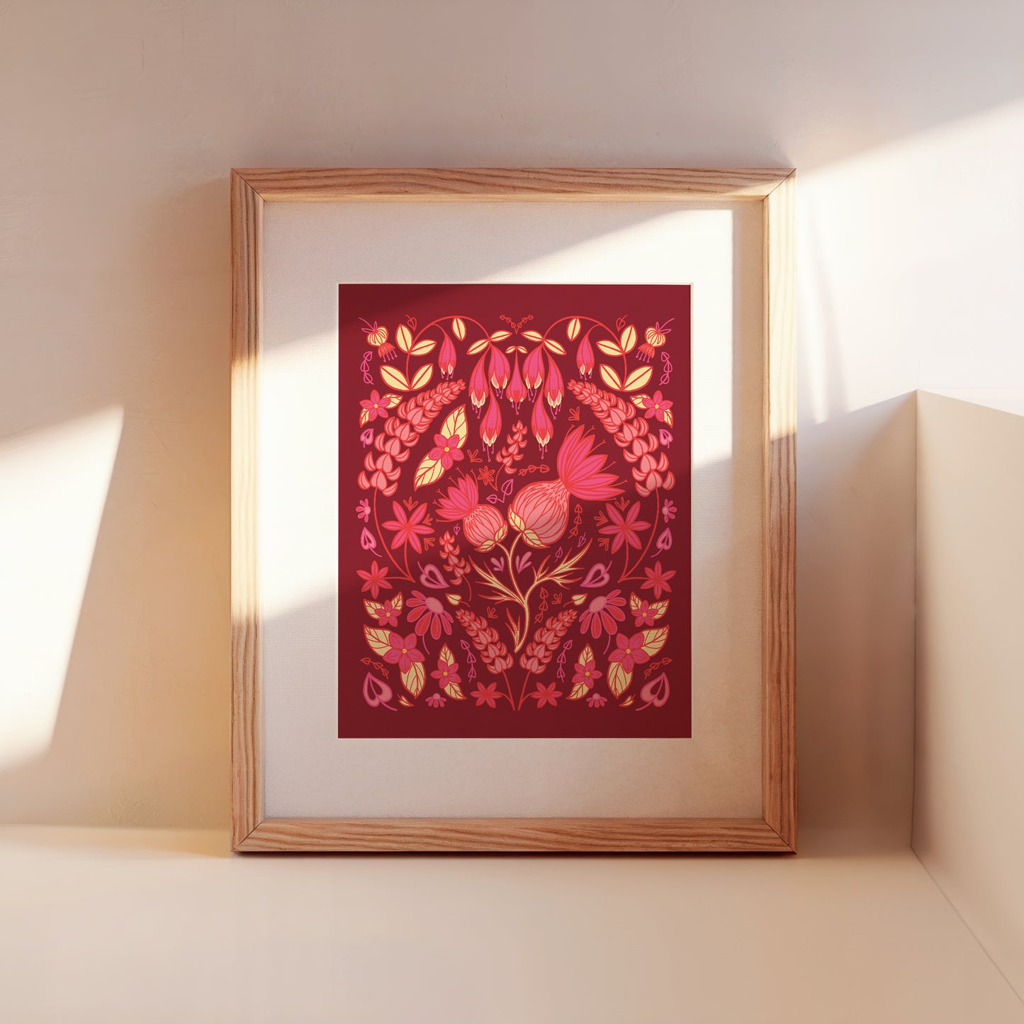 A framed burgundy and pink thistle folk floral leans against a wall.