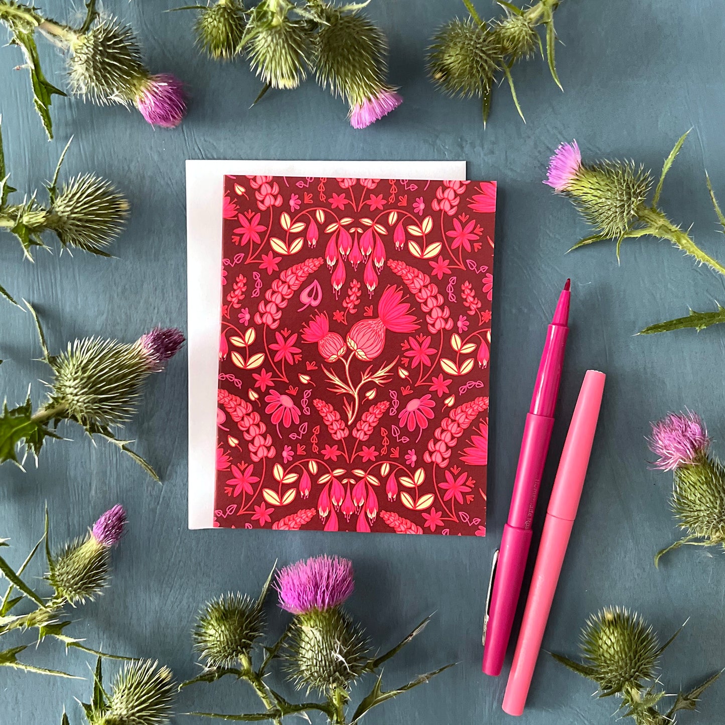 A dark pink greeting card is covered in a folk-style pattern of pink thistles, lupines, haskap berries and mayflowers, with creamy yellow leafy accents. The card is flanked by thistles and pink pens.
