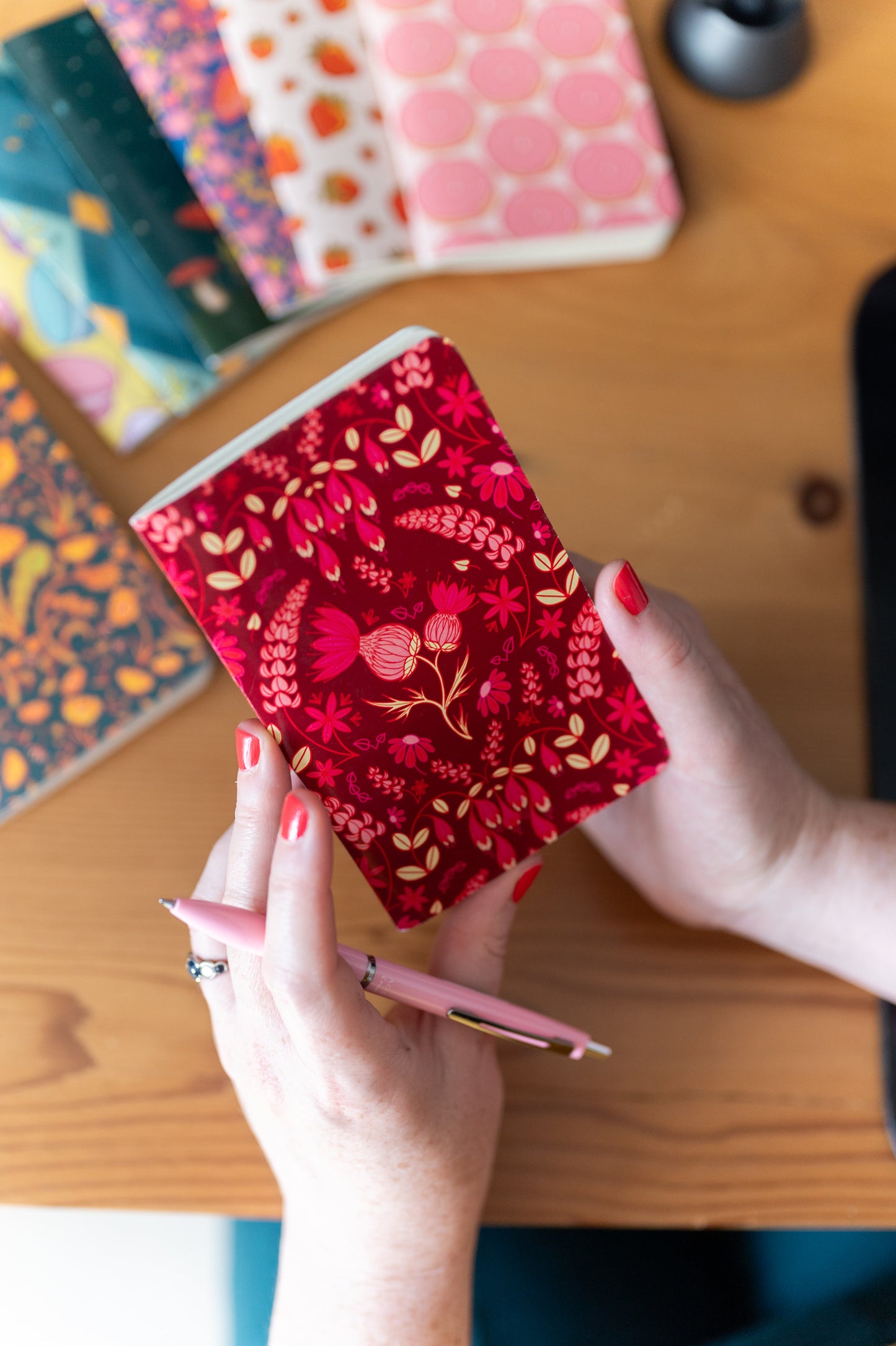 A person holds a red notebook patterned with thistles, lupines and coneflowers. In the background are other colourful notebooks.