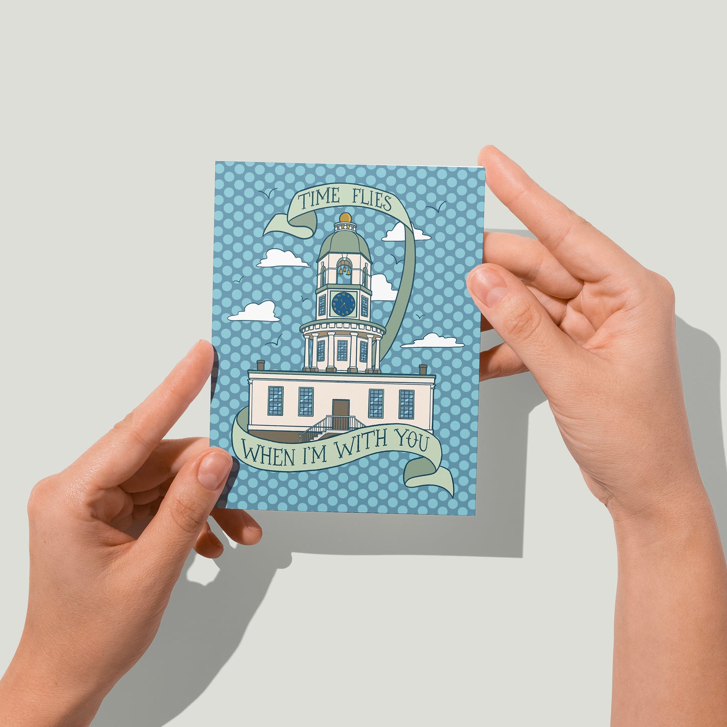 A blue polka dot greeting card features an illustration of the Halifax town clock with scattered clouds and seagulls. A hand-lettered banner reads Time Flies when I'm with You. A pair of hands holds the card up in front of the camera.