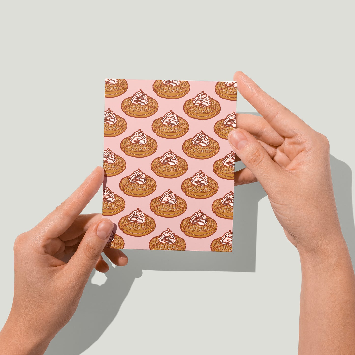 A pink greeting card is patterned with Belgian waffles topped with whipped cream. A pair of hands holds the card up in front of the camera.