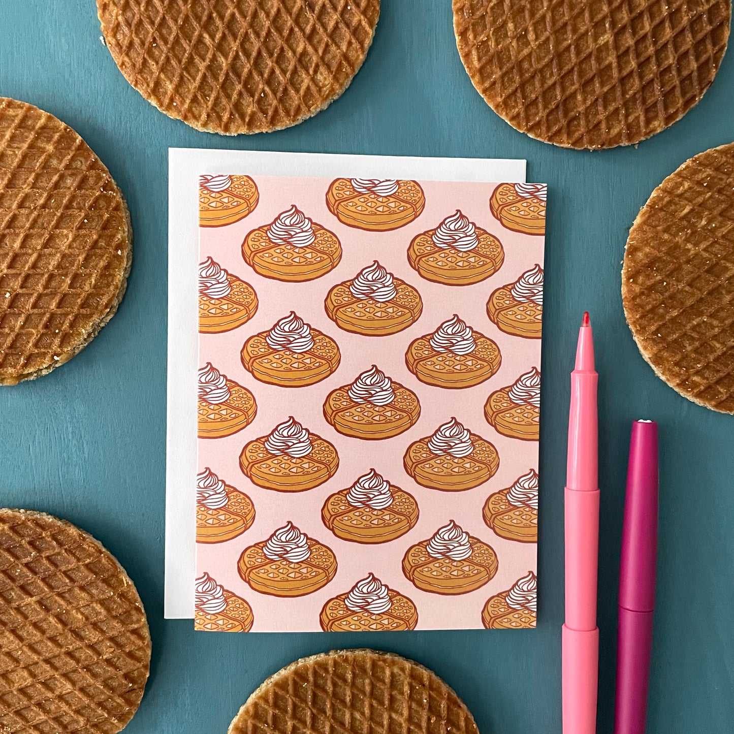 A pink greeting card patterned with Belgian waffles topped with whipped cream is surrounded by stroopwafels and felt-tip pens on a blue background.