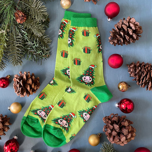 A pair of light green socks patterned with animatronic Christmas trees and gifts lies flat among greenery, pinecones and Christmas tree ornaments.