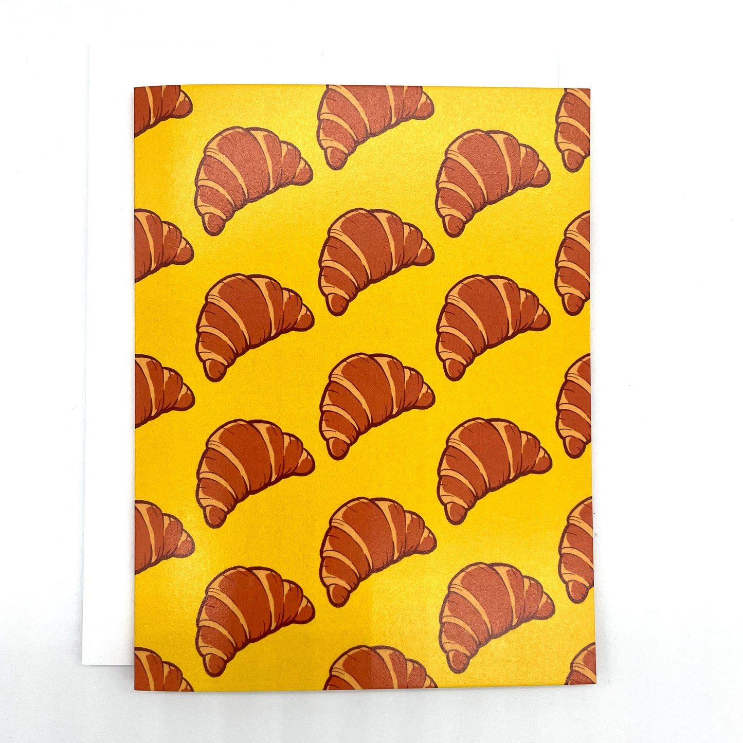 A yellow greeting card is patterned with drawings of croissants in diagonal lines. The card sits against a white envelope on a white background.