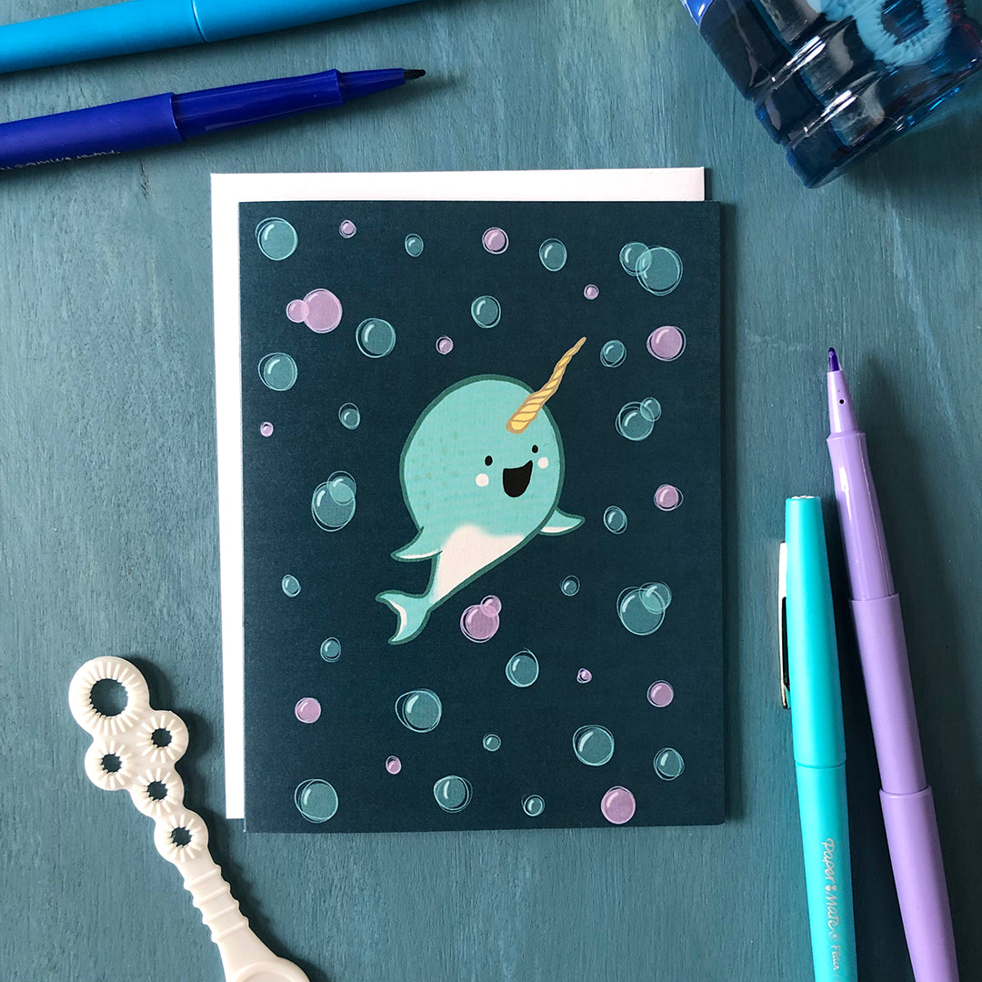 A dark blue greeting card shows an illustration of a cute, happy light blue baby narwhal surrounded by blue and purple bubbles. Around the card are blue and purple pens, a bubble wand, and a bottle of bubble liquid against a bue wooden background.