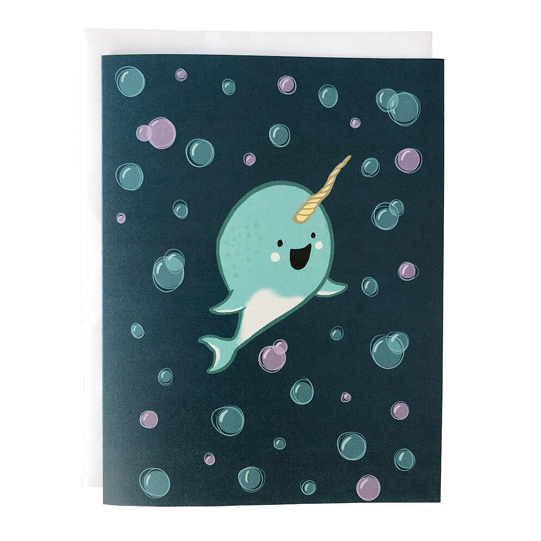 A dark blue greeting card shows an illustration of a cute, happy light blue baby narwhal surrounded by blue and purple bubbles.The card sits against a white envelope on a white background