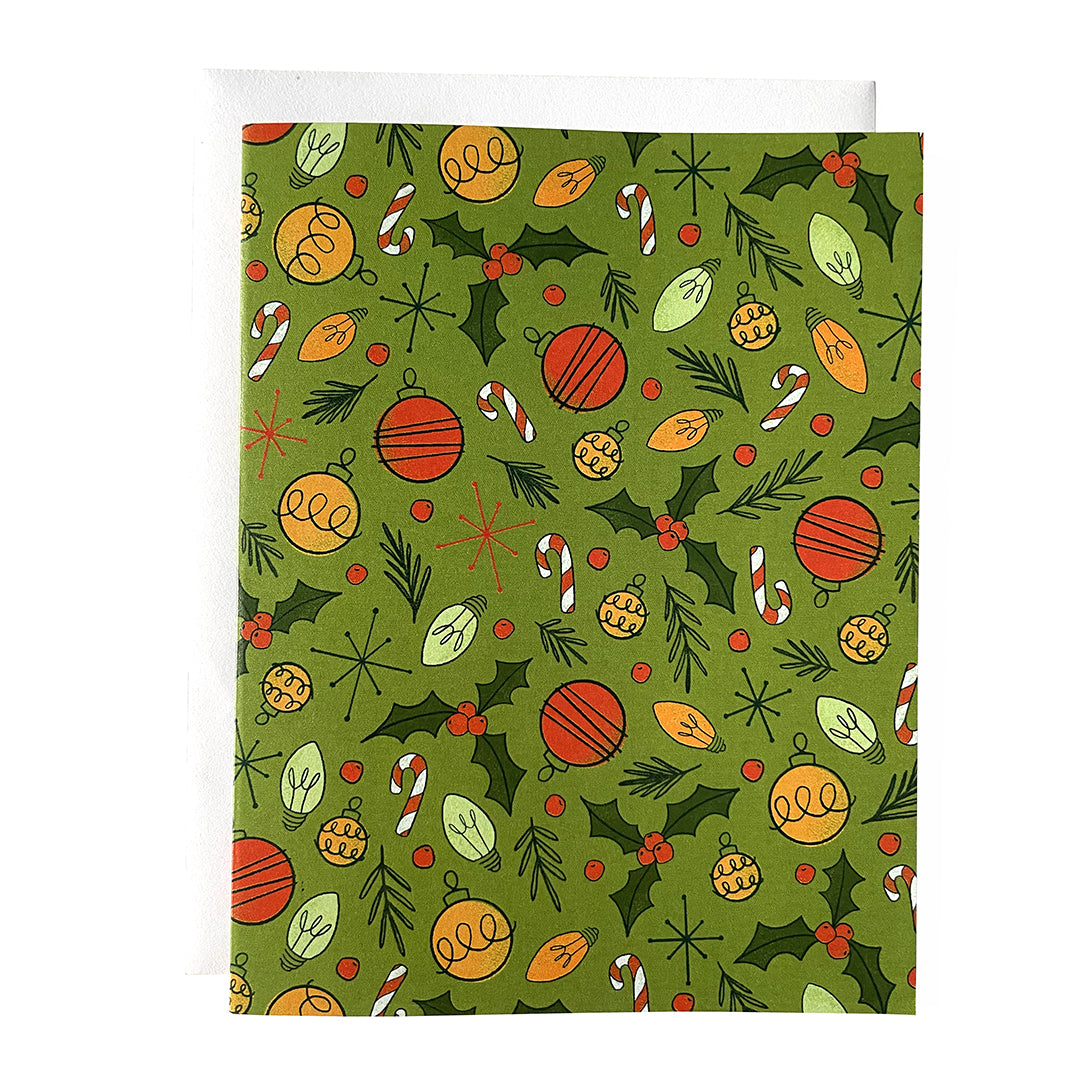 A green greeting card is patterned with midcentury-inspired ornaments, holly, candy canes and Christmas lights. The card is against a white envelope on a white background.