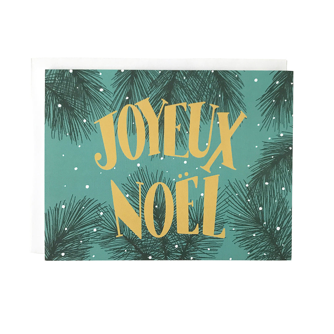 A teal greeting card features the words Joyeux Noël in midcentury-inspired lettering against a background of snowy evergreen branches. The card sits against a white envelope on a white background.