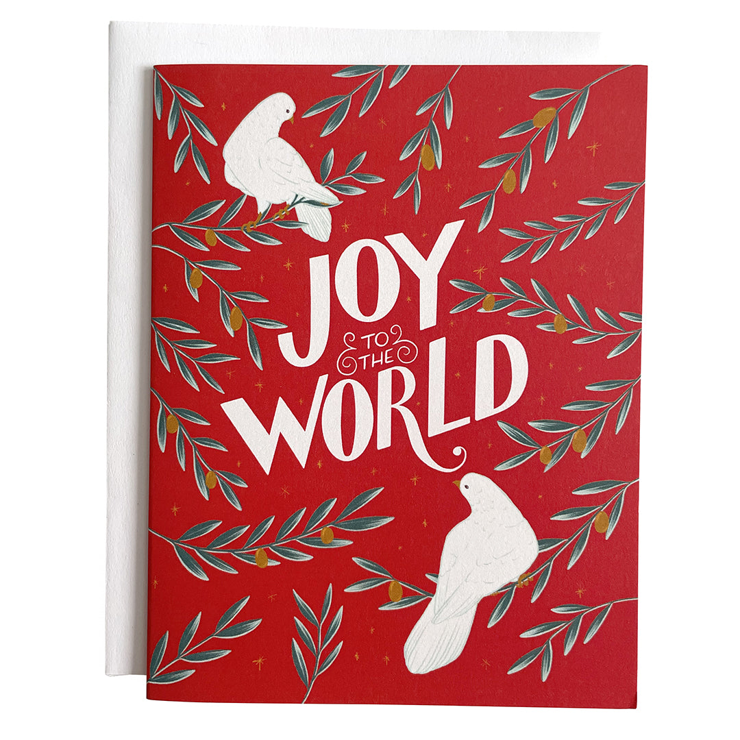 A red greeting card shows two white doves, branches of golden olives, and the hand-lettered words Joy to the World in white. The card is against a white envelope on a white background.