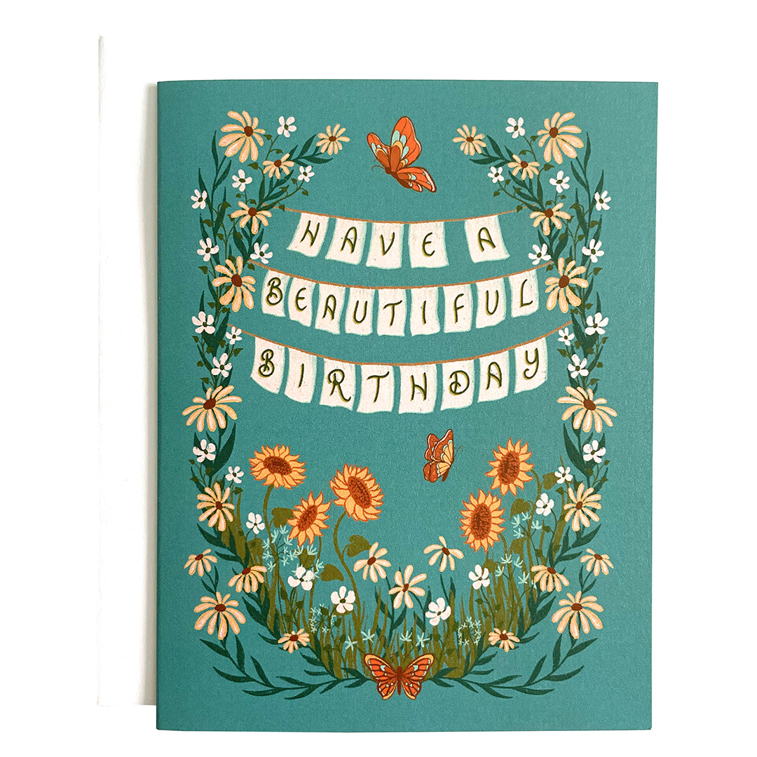 A blue cottagecore greeting card tells you to have a beautiful birthday. The words are written on banners strung between sunflowers and daisies, and orange butterflies appear above and below the letters. The card is on a white envelope on a white background.
