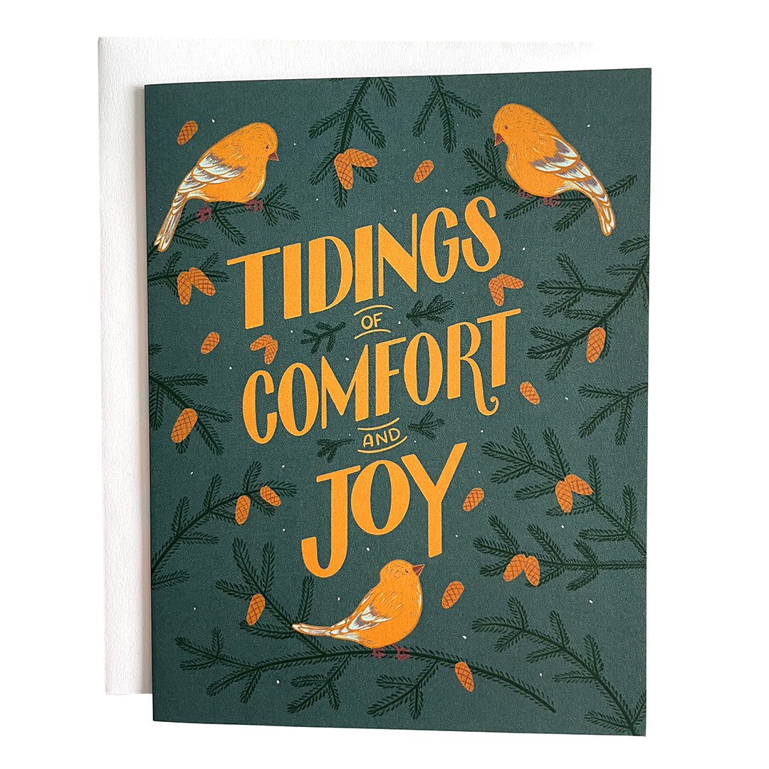 A greeting card features yellow hand-lettering reading Tidings of Comfort and Joy on a blue background. There are painted goldfinches and pinecone-laden branches around the words. The card is on a white envelope on a white background.