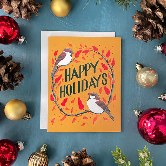 A yellow greeting card features the words Happy Holidays in hand lettering, surrounded by sparrows and staghorn sumac. The card is surrounded by red and gold ornaments, pinecones, and greenery.