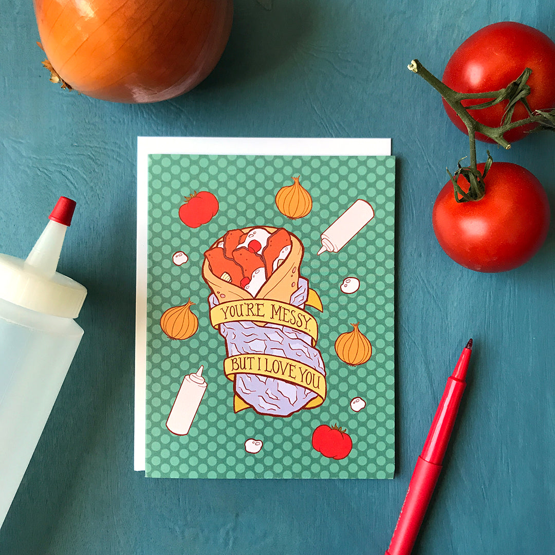 A green polka dot greeting card features an illustration of a donair wrapped in foil, with squeeze bottles, onions and tomatoes all around it and a banner that reads You're Messy But I Love You. Around the card are tomatoes, an onion and a squeeze bottle.