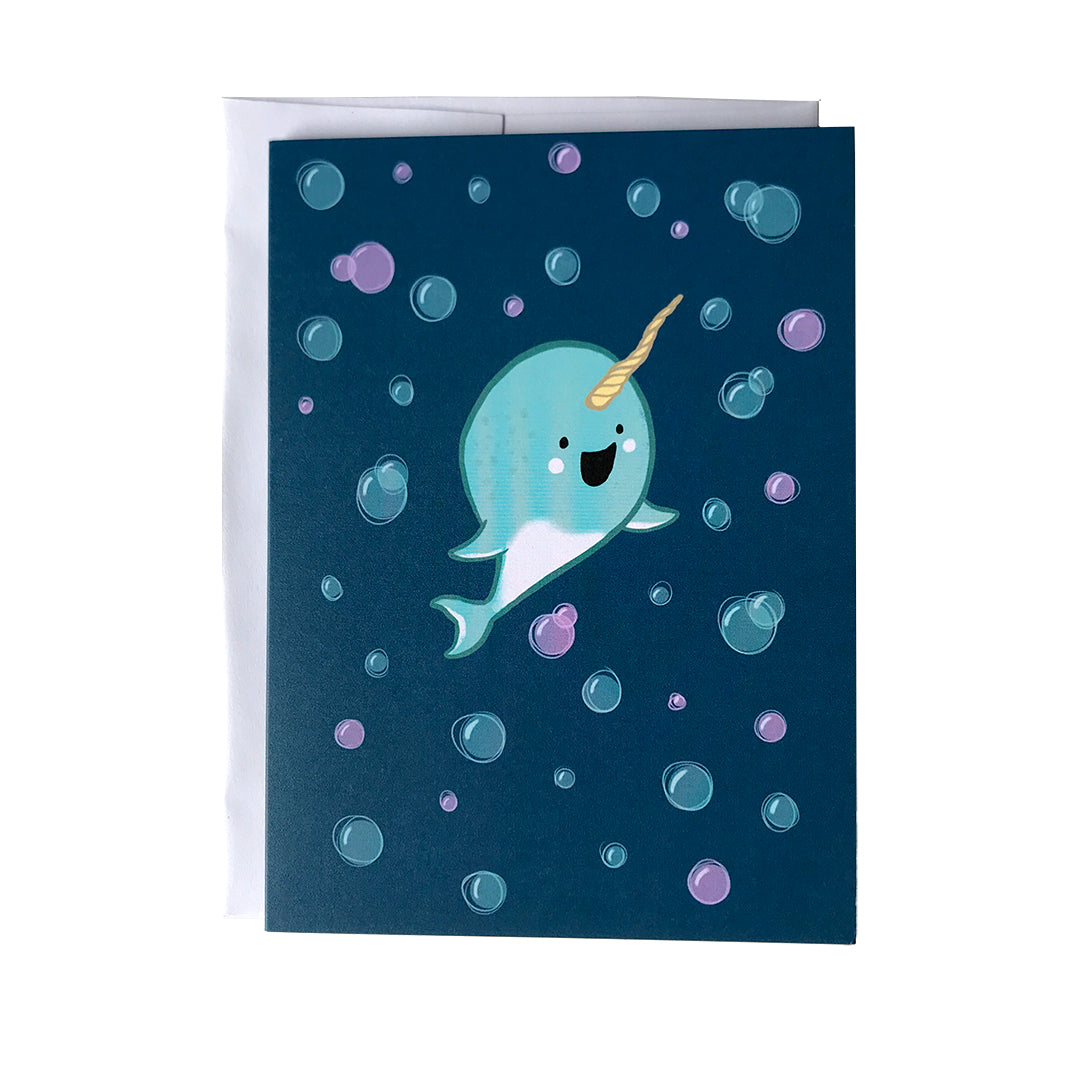 A dark blue greeting card shows an illustration of a cute, happy light blue baby narwhal surrounded by blue and purple bubbles.The card sits against a white envelope on a white background.