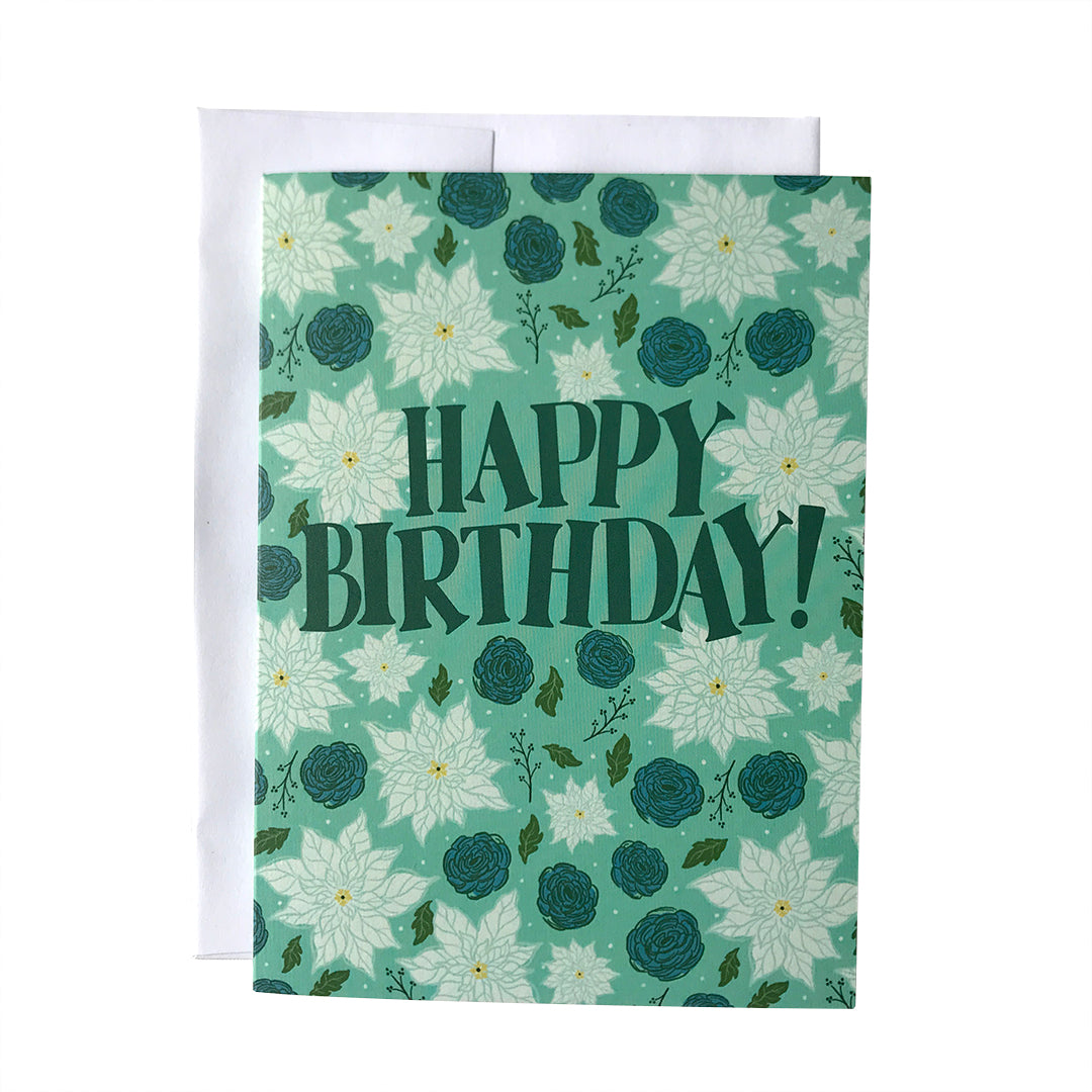 A blue greeting card featuring the words Happy Birthday! in midcentury-inspired dark blue capital letters. Behind and all around the letters are white poinsettias, blue ranunculus flowers, green leaves and black branches. The card sits against a white envelope on a white background.