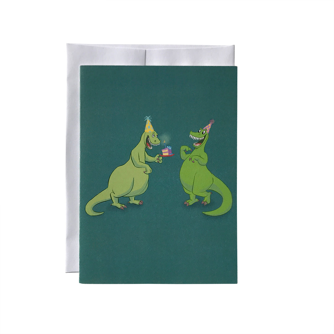 A blue-green greeting card shows an illustration of two grinning green dinosaurs in party hats. One offers a slice of cake to the other. The card sits against a white envelope on a white background.