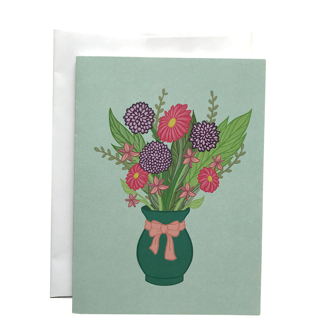 A pastel mint greeting card features an illustration of a bouquet in a green vase with a pink bow around it. The bouquet includes purple and pink flowers and greenery in various shades. The card sits against a white envelope on a white background.