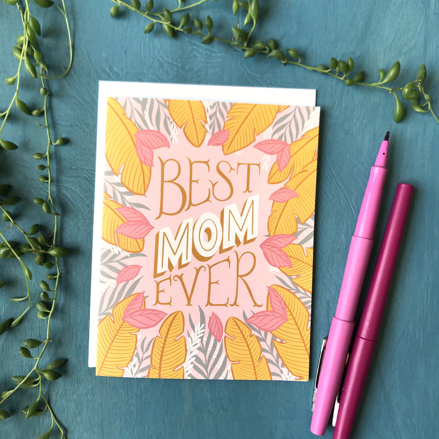 A pink card covered in golden yellow, white and grey tropical leaves with the lettered phrase "Best Mom Ever" rests on a blue background beside 2 pink pens. Buds from a string of pearls plant surround it.