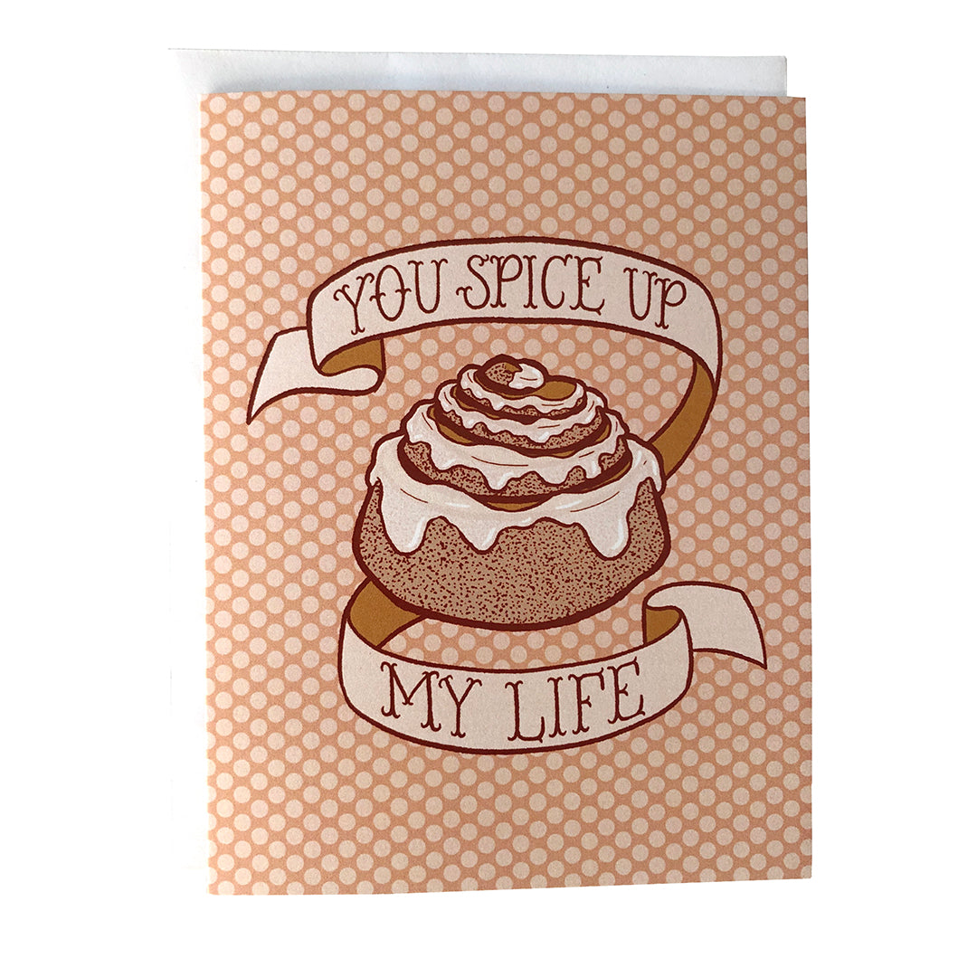 A peach polka dot greeting card bears an illustration of a cinnamon bun dripping with frosting and a banner that reads you spice up my life in hand lettering. The card sits against a white envelope on a white background.