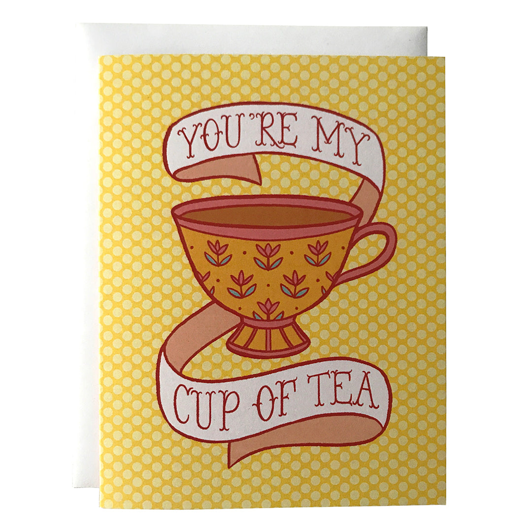 A yellow polka dot greeting card features an illustration of a golden teacup with yellow flowers, full of strong tea. Around the teacup is a banner that reads You're my cup of tea in pink letters. The card sits against a white envelope on a white background.