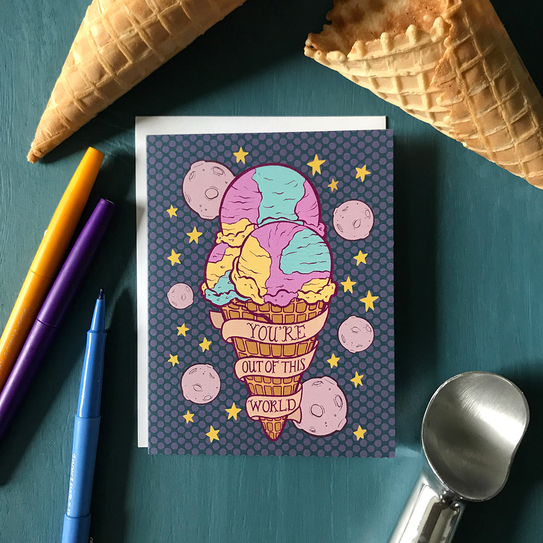 A purple greeting card featuring a large Moon Mist ice cream cone, purple moons, yellow stars and a banner that reads you're out of this world rests on a blue background. Around it are colourful pens, an ice cream scoop and waffle cones.