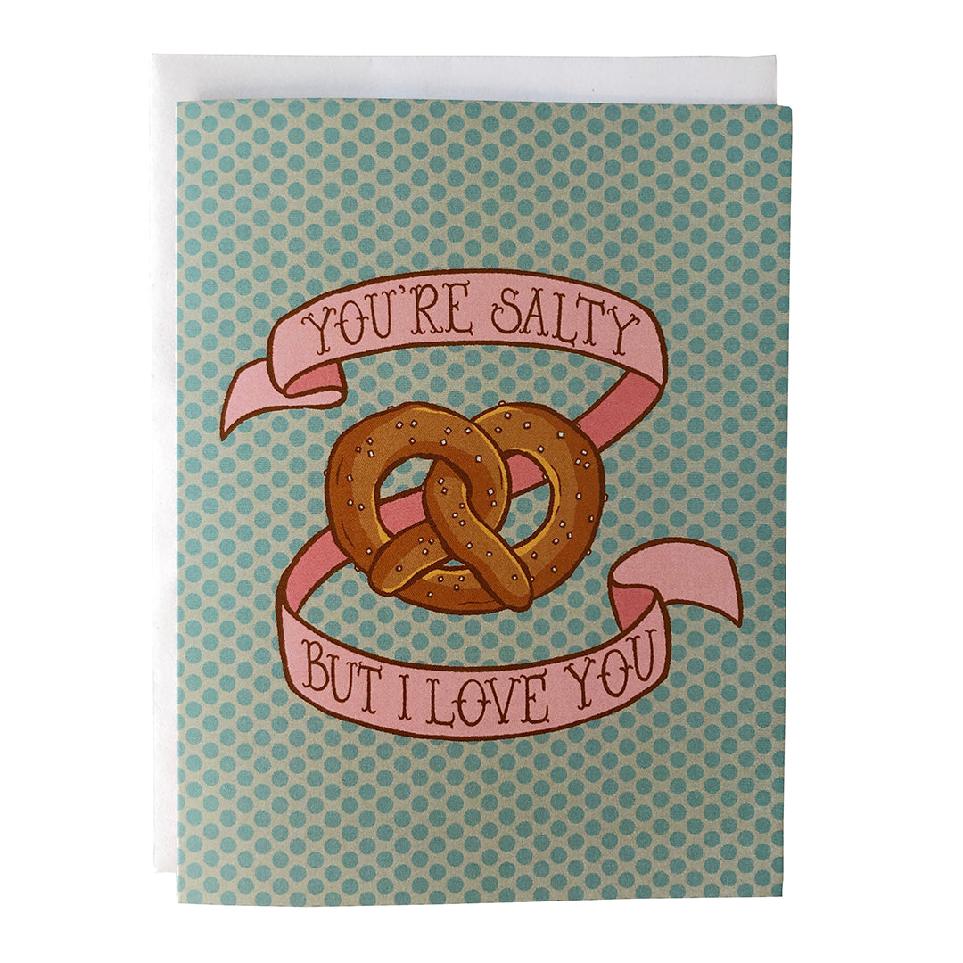 A pastel polka dot greeting card shows an illustration of a soft salted pretzel surrounded by a pink banner that reads you're salty but I love you in hand lettering. The card sits against a white envelope on a white background.