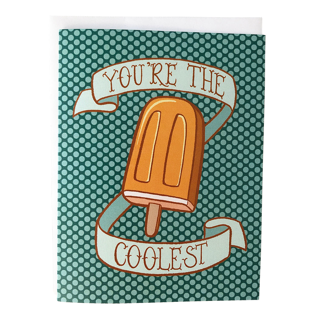 A turquoise polka dot greeting card shows an illustration of a creamy orange popsicle surrounded by a banner that reads you're the coolest in hand lettering. The card sits against a white envelope on a white background.