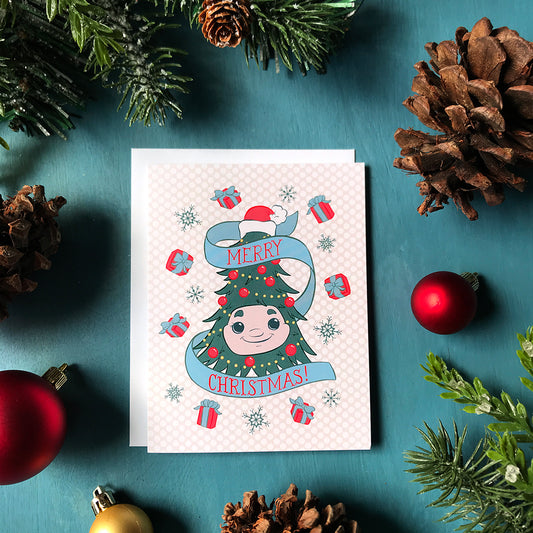 A Christmas card featuring an illustration of Woody the Christmas Tree, some red gifts with blue bows and blue snowflakes on a pale pink polka dot background. The card sits on a blue background surrounded by red and gold ornaments, pinecones and faux evergreen branches.