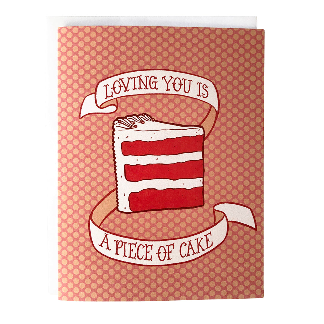 A pink polka dot greeting card shows an illustration of a triple-layer slice of red velvet cake and a banner that reads loving you is a piece of cake in hand lettering. The card sits against a white envelope on a white background.