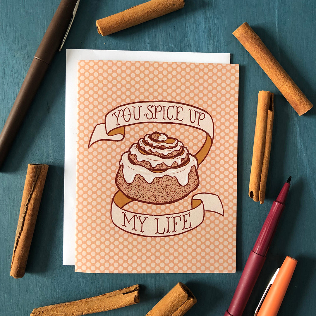 A peach polka dot greeting card bears an illustration of a cinnamon bun dripping with frosting and a banner that reads you spice up my life in hand lettering. The card is surrounded by brown, raspberry and orange pens and several cinnamon sticks on a blue wooden background.